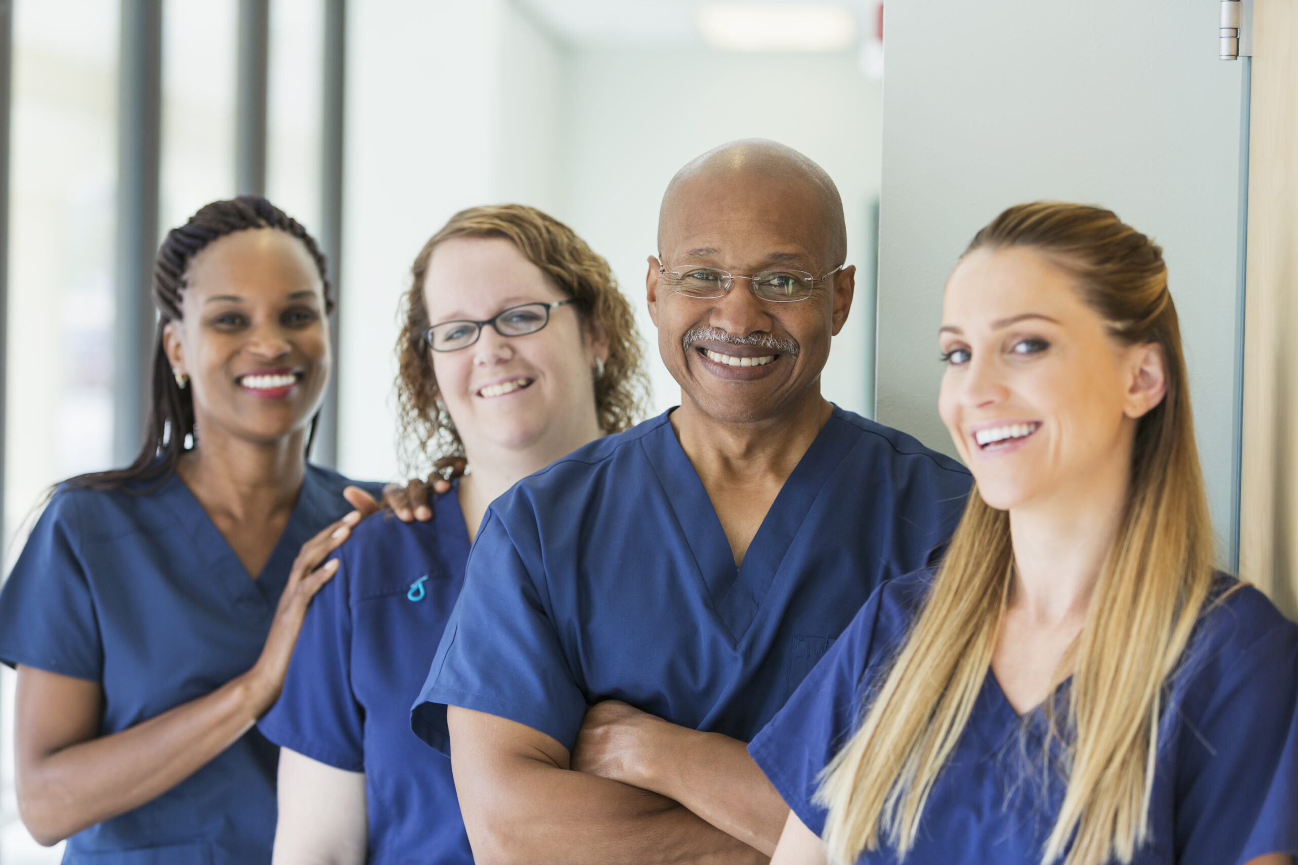 A team of four multi-ethnic medical professionals standing in a corridor, wearing blue scrubs, smiling at the camera. The focus is on the African-American senior man, in his 60s.